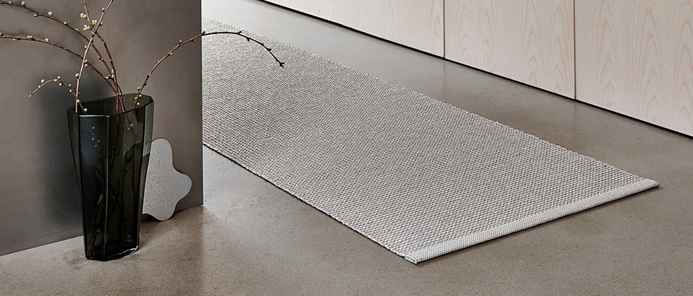 A minimalist interior featuring the Pappelina Emm linen rug, placed along a polished concrete floor near light wood paneling. The scene includes a dark glass vase with branches and a sculptural object.